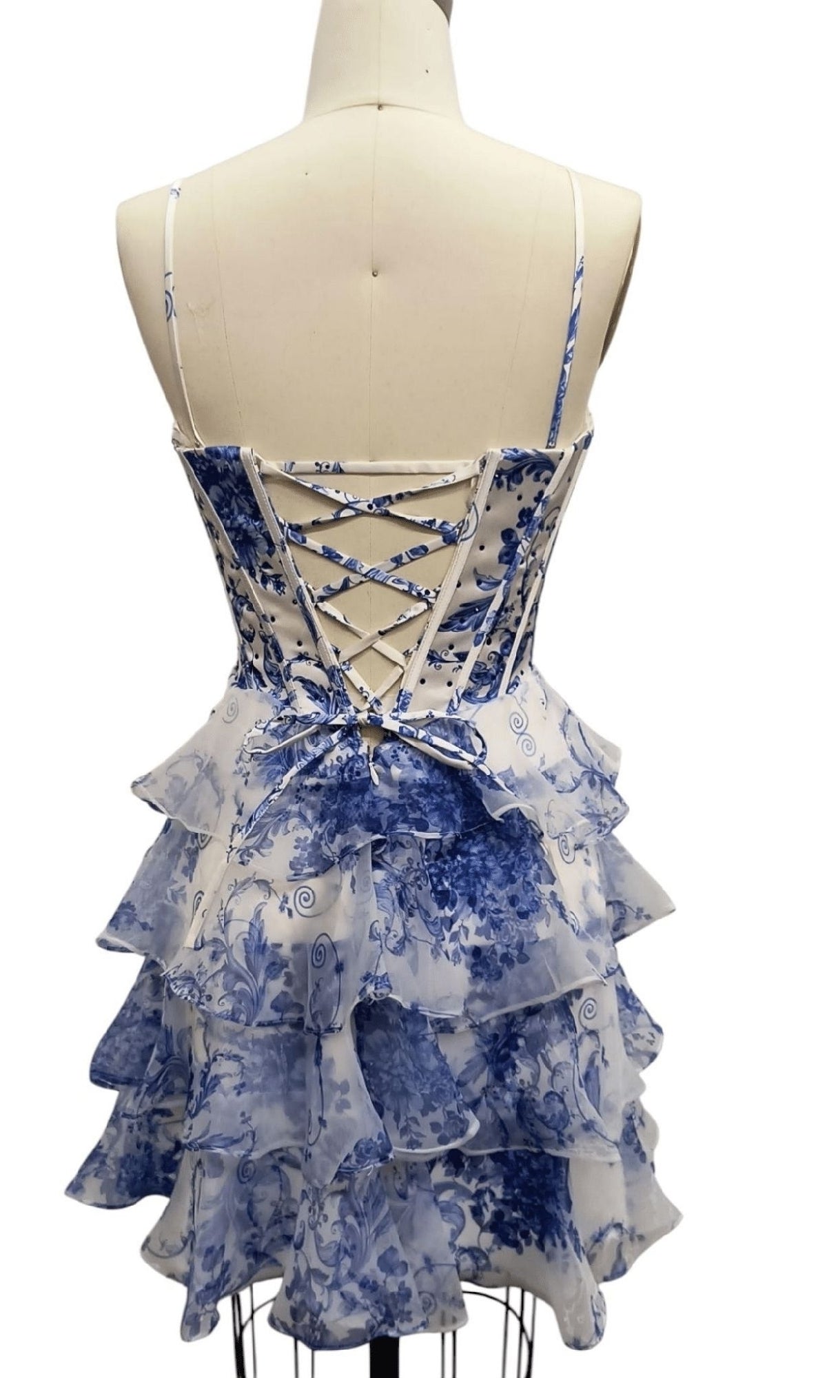 A mannequin showcases the Short Print Corset Hoco Dress: 12613, a blue printed homecoming dress with layered ruffles and a lace-up back, combining vintage charm and playful elegance.